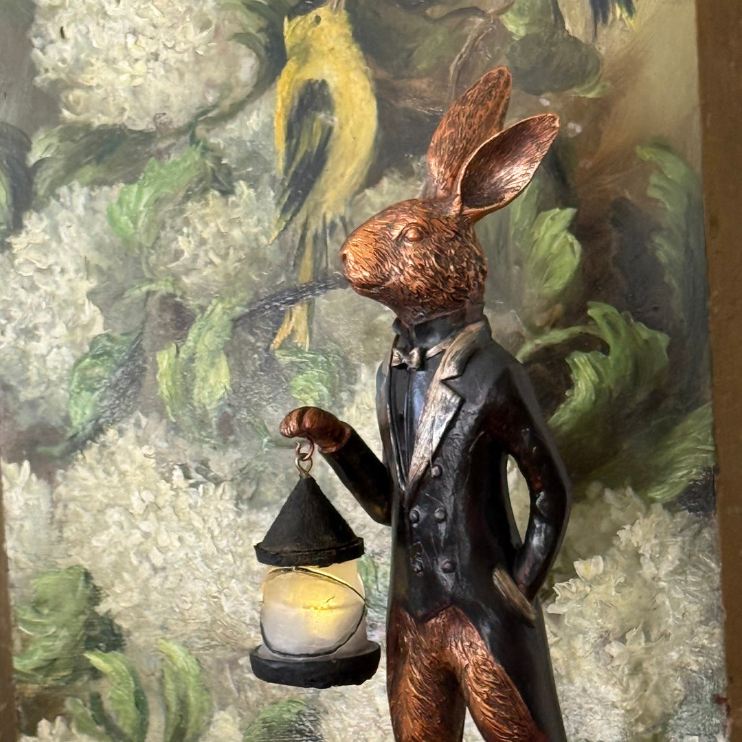 Close-up of a bronze hare figurine carrying a small LED lantern, dressed in a vintage-style coat and bow tie, with an oil painting featuring hydrangeas and birds in the background