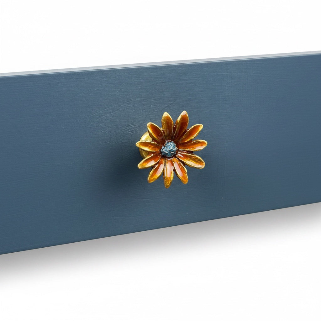 A close-up of a decorative flower-shaped cabinet knob with orange and gold petals and a dark blue center, mounted on a blue painted wooden surface.