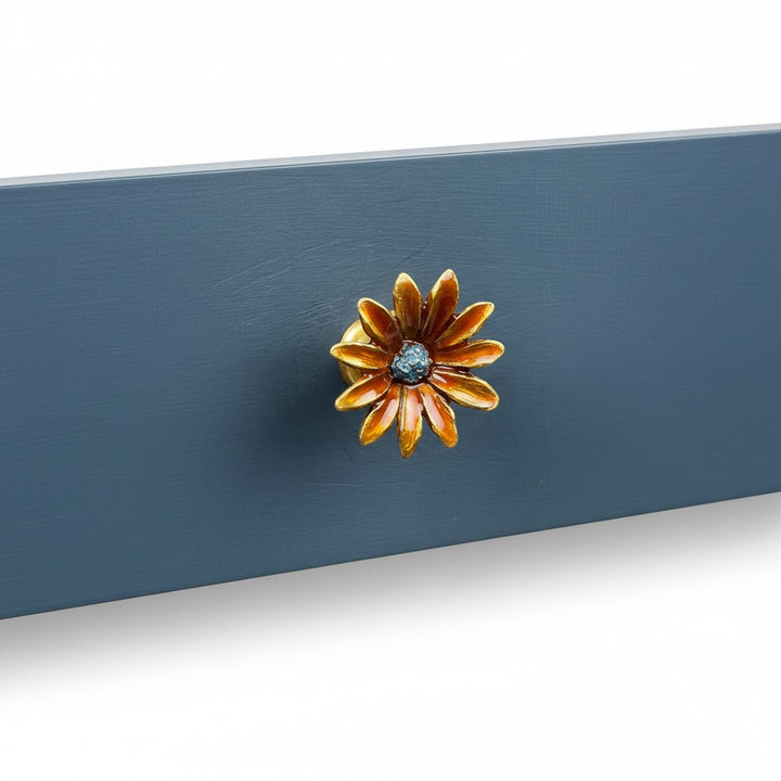 A close-up of a decorative flower-shaped cabinet knob with orange and gold petals and a dark blue center, mounted on a blue painted wooden surface.