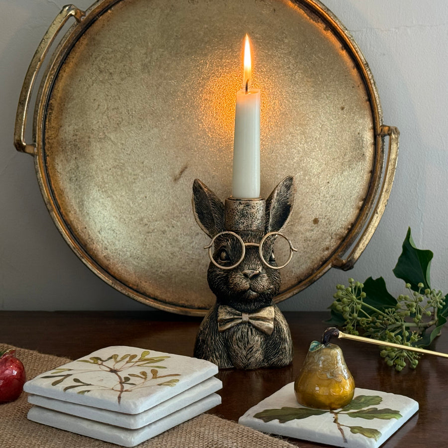 Decorative rabbit figurine with glasses and a candle, placed on a table with coasters and a plant.
