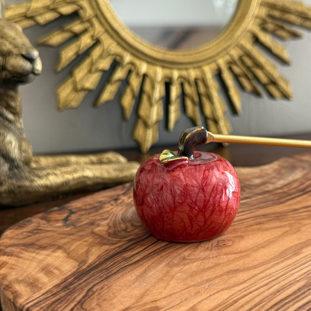 Red apple candle snuffer with a gold stem on a wooden surface in front of a decorative mirror.
