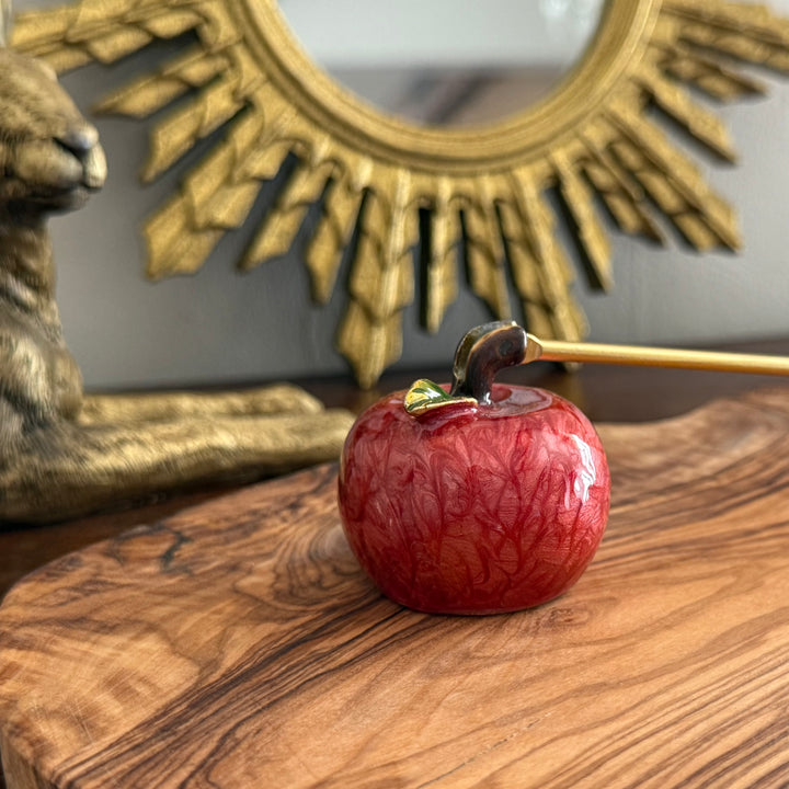 Red apple candle snuffer with a gold stem on a wooden surface in front of a decorative mirror.