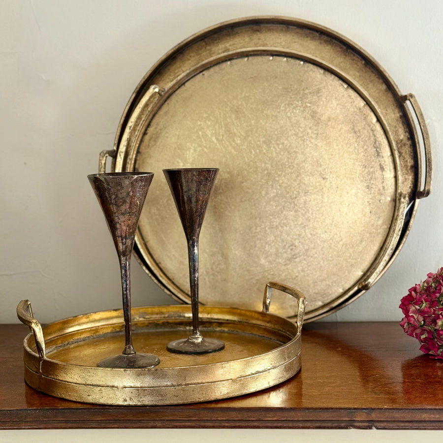 gold metal tray with handles on a dark wood table, holding 2 tall pewter glasses with 2 larger gold trays nesting behind