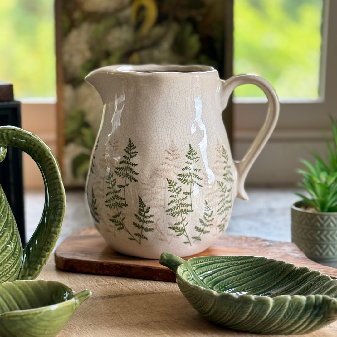 White ceramic pitcher with fern design on a wooden surface, surrounded by green leaf-shaped bowls.