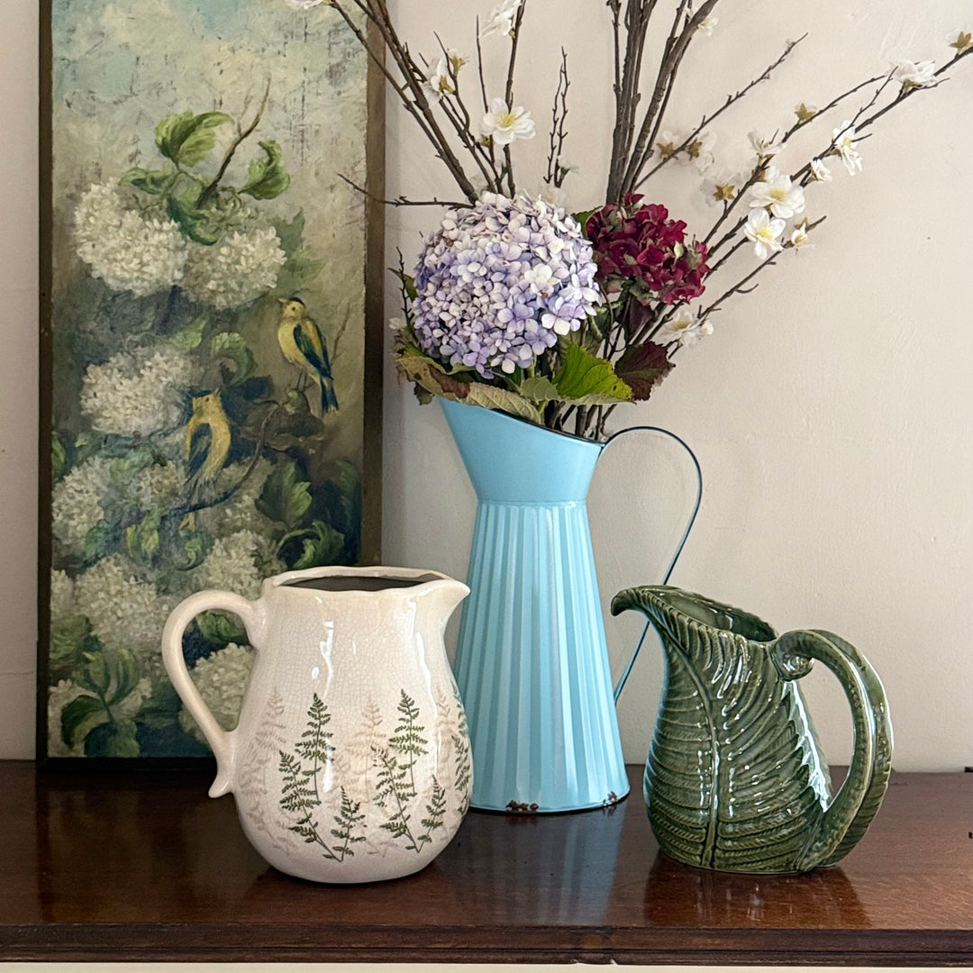 Three pitchers on a wooden surface with a floral arrangement and painting in the background.