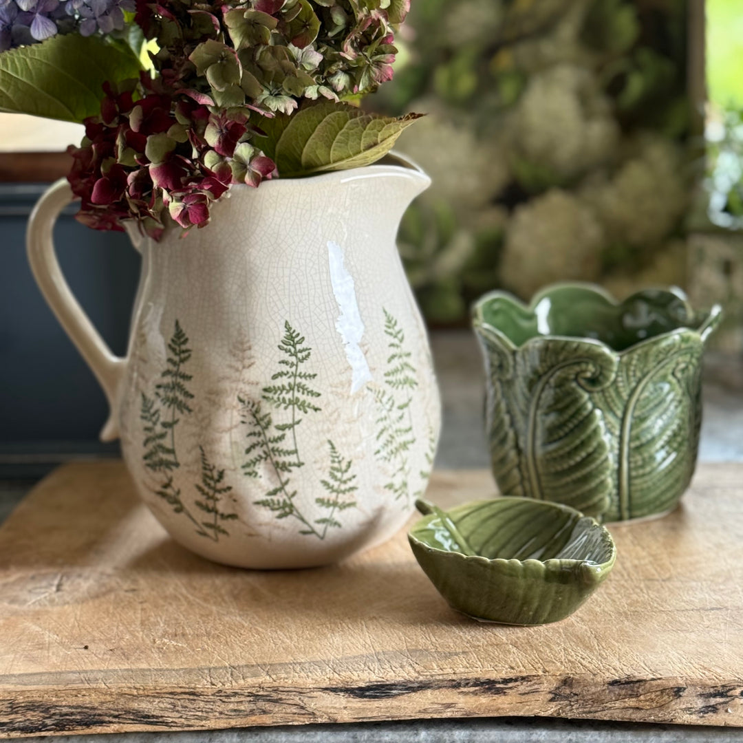 White pitcher with green leaf patterns on a wooden surface with a green bowl and flowers.
