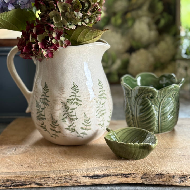 White pitcher with green leaf patterns on a wooden surface with a green bowl and flowers.