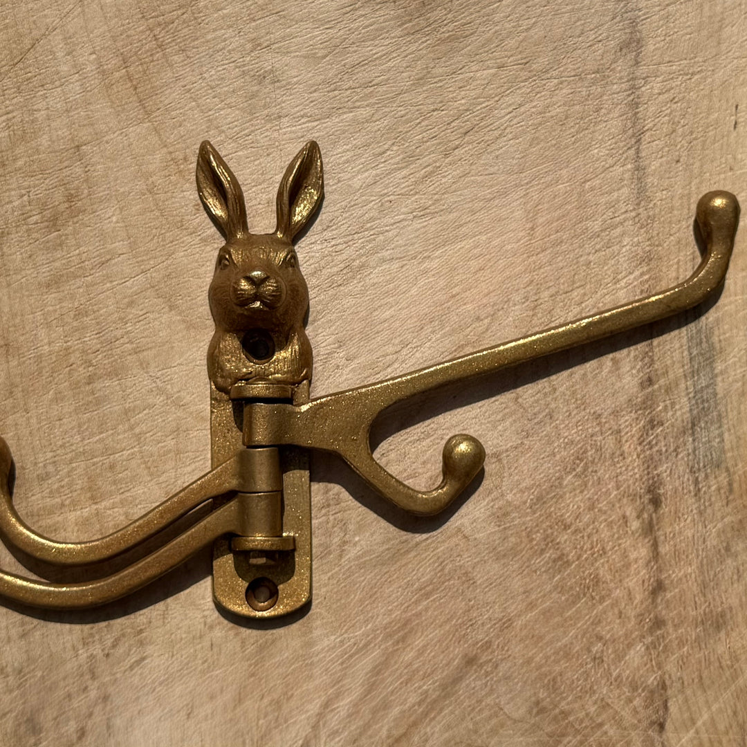 An antique gold rabbit-shaped coat rack hook with two curved hooks, shown mounted on a light wood background.