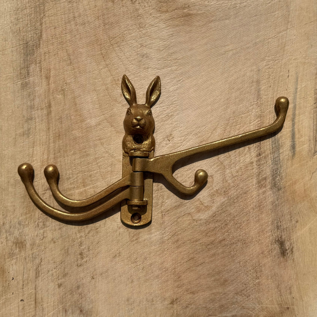 A vintage-style gold rabbit door hook with three curved hooks, mounted on a rustic wooden surface.