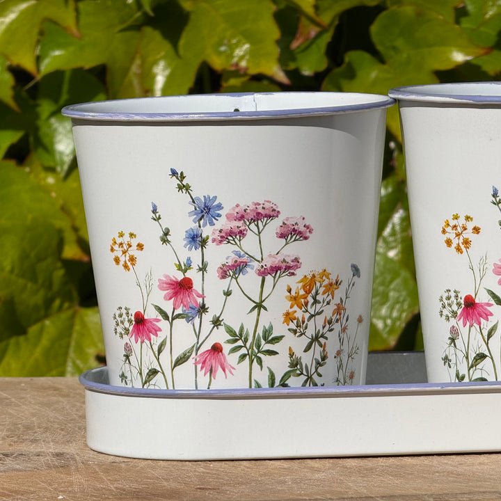 Two white enamel mugs with floral designs on a wooden surface with green leaves in the background.
