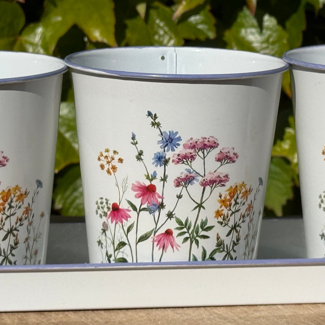 Three white enamel pots with floral designs on a wooden surface with green foliage in the background.