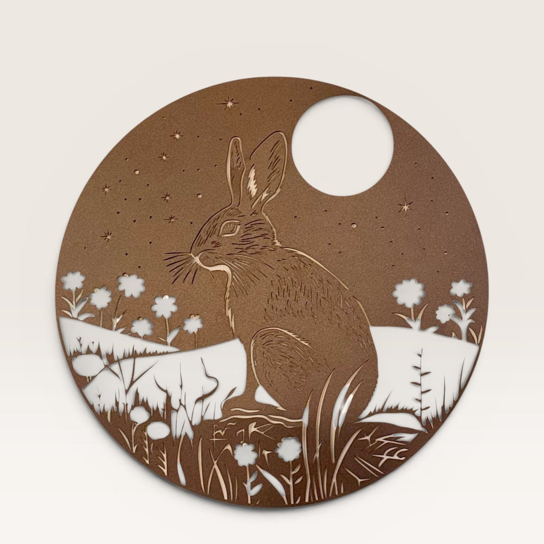 A circular brown metal cutout featuring an illustration of a hare sitting in a field of flowers under a full moon and starry sky, displayed against a plain off-white background.