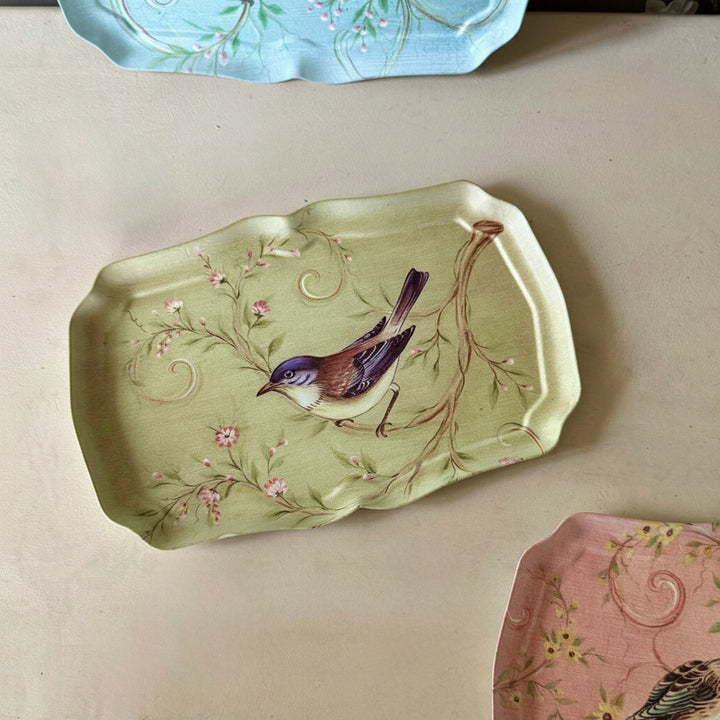A rectangular metal  tray with scalloped edges, featuring a painted bird perched on a flowering branch over a pale yellow-green background. Partially visible at the top and bottom are other decorative trays with floral patterns.