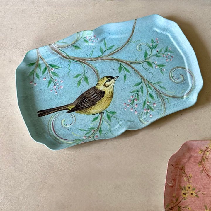 A light blue metal trinket
 tray with a hand-painted yellow bird perched on a branch with green leaves and small pink blossoms, displayed on a neutral beige surface.