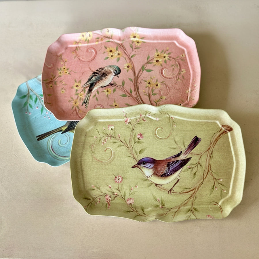Three decorative serving trays with scalloped edges, featuring bird and floral designs. One tray is light pink, one is pale green, and one is light blue, arranged on a beige surface.