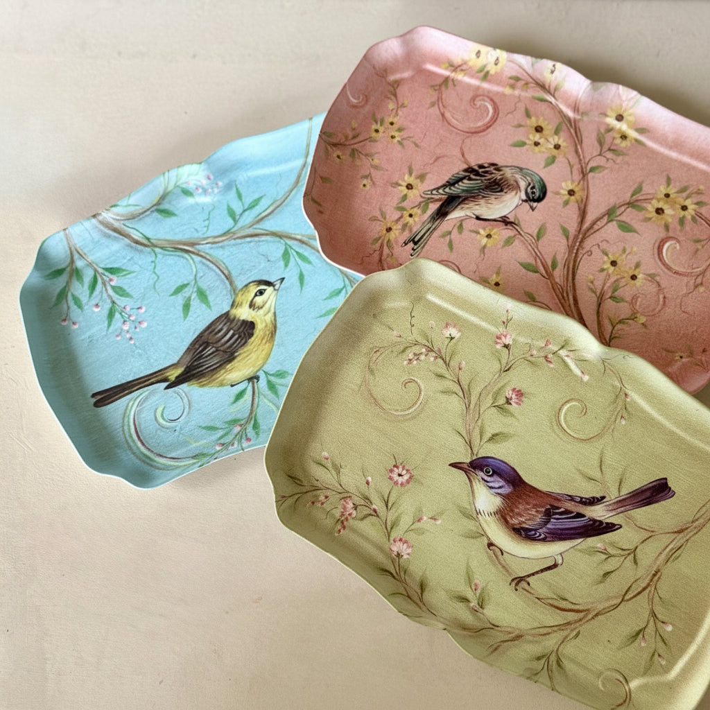 Three decorative trinket trays with scalloped edges, each featuring a painted bird and floral branch pattern. One tray is light blue, one is dusty pink, and one is pale olive green, arranged on a light beige surface.