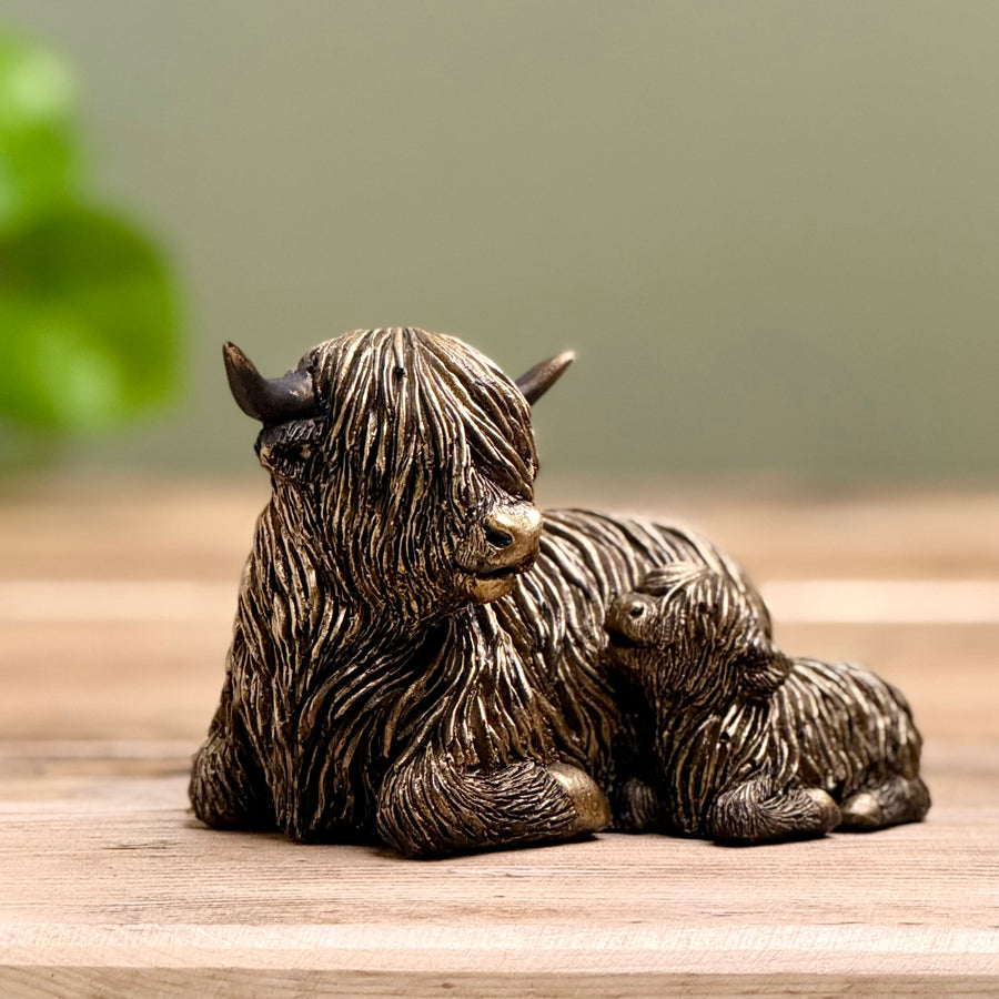 A bronze-finished figurine of a highland cow with its calf, resting on a wooden surface with a soft-focus green background.