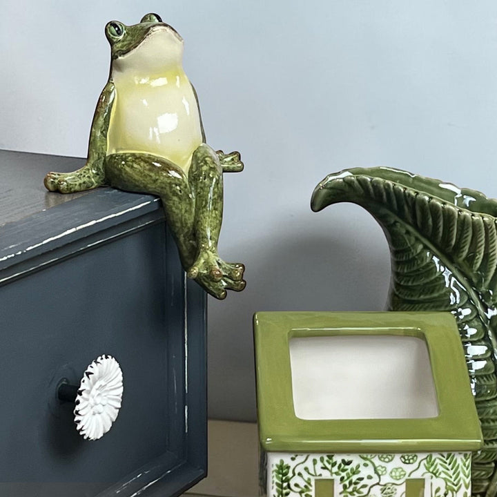 Green polyresin frog shelf sitter with crossed legs perched on a grey shelf edge with a green fern jug and planter on a table