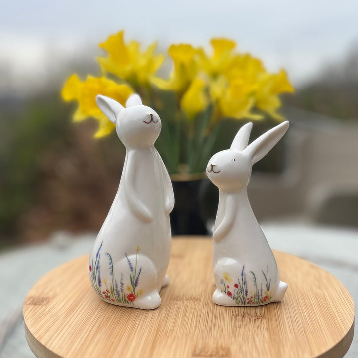 Two white ceramic rabbit figurines with floral designs on a wooden surface, with yellow flowers in the background.
