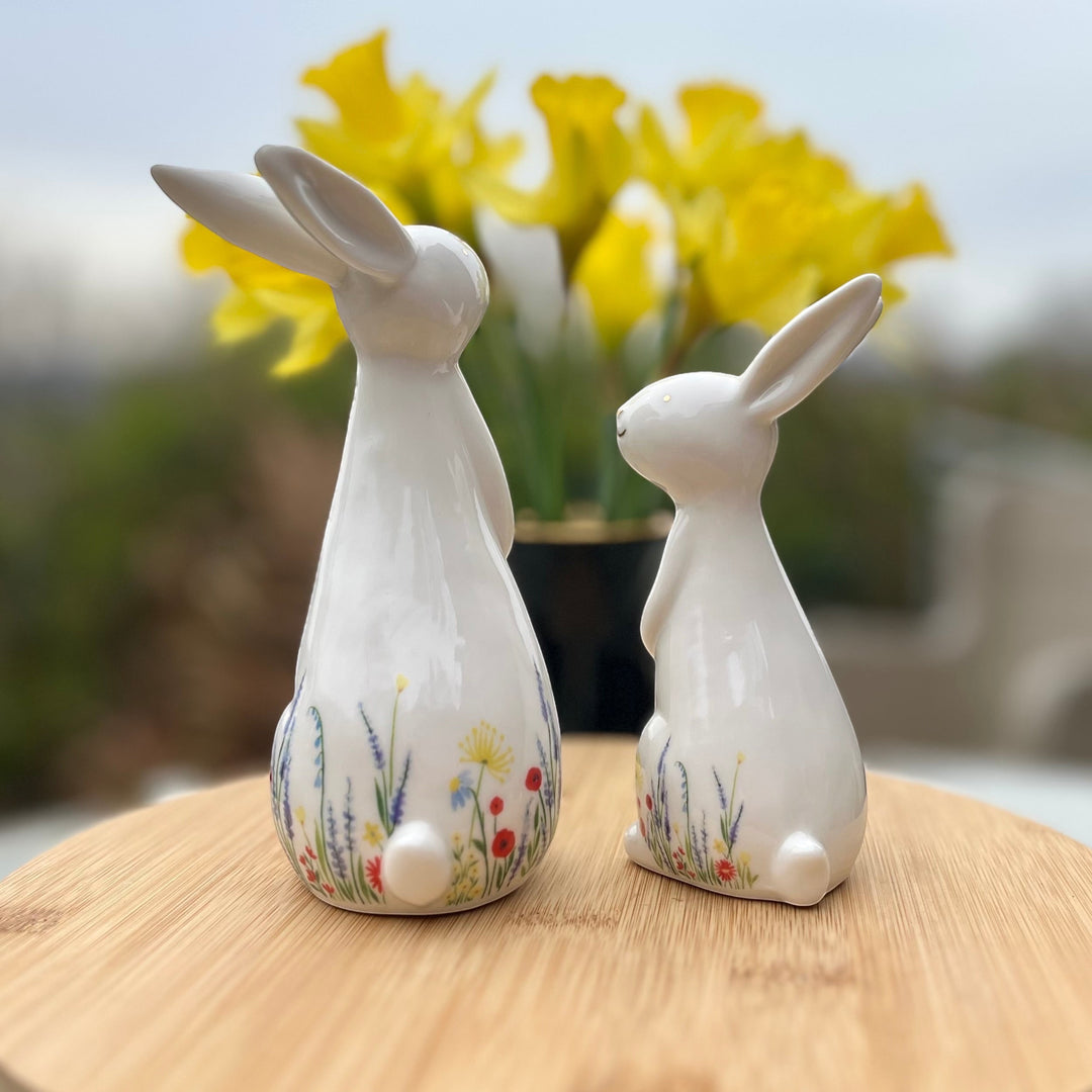 Two ceramic rabbit figurines with floral designs on a wooden surface with a blurred background of yellow flowers.