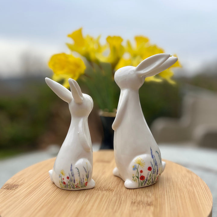 Two white ceramic rabbit figurines with floral designs on a wooden surface with a blurred background of yellow flowers.