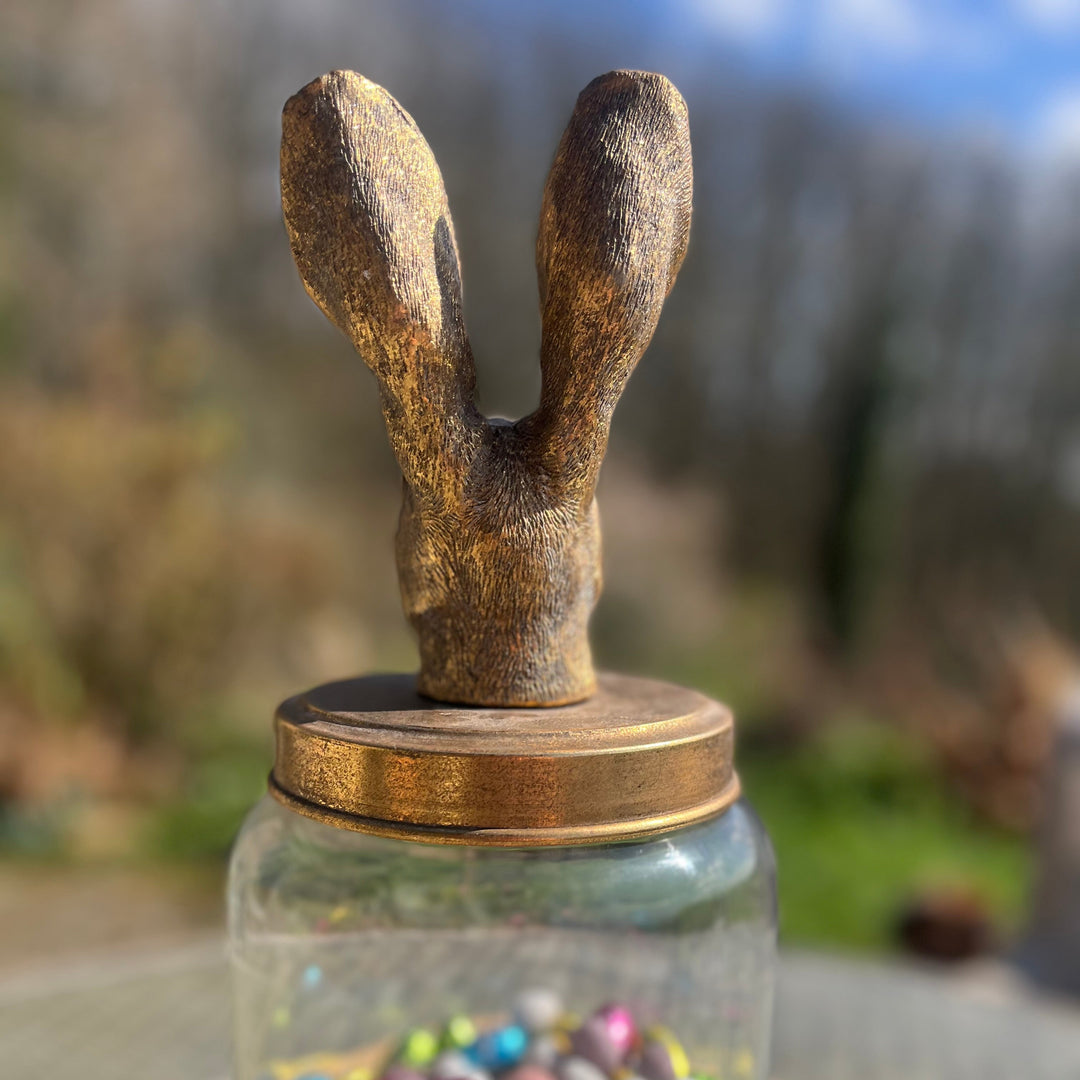 A clear glass jar filled with colorful pastel chocolate eggs, topped with a rustic gold colour bunny head lid, displayed on a wooden stand outdoors.