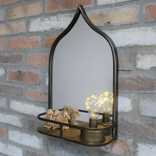 An arched wall mirror with a dark metal frame and shelf, styled with small figurines and glowing light bulbs on a brick wall background.