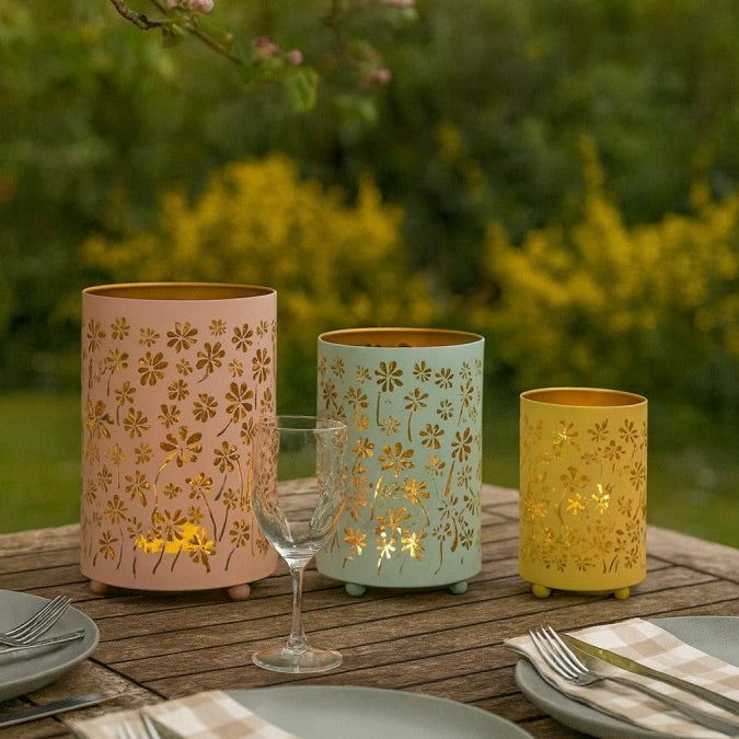Three metal flower lantern candle holders in pink, mint green, and yellow are arranged on a wooden outdoor table with plates, a wine glass, and a blurred garden background.