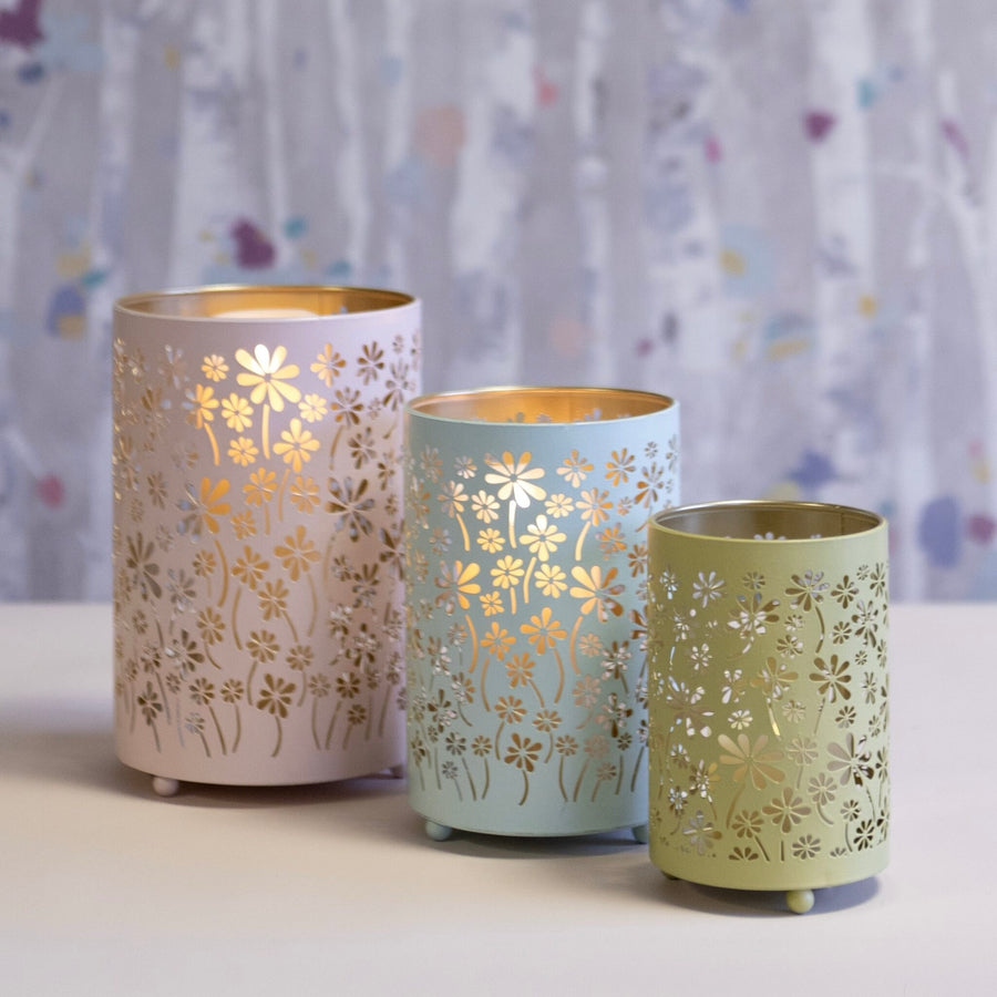 Three floral metal candle holders in pastel pink, light blue, and sage green, each with cutout flower patterns,  displayed on a white surface against a softly blurred pastel background.