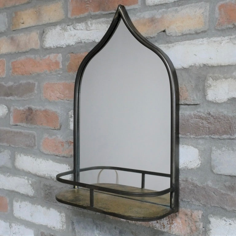An arched wall mirror with a dark metal frame and a matching shelf, mounted on a rustic brick wall.