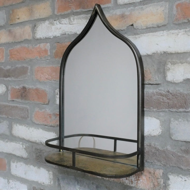 An arched wall mirror with a dark metal frame and a matching shelf, mounted on a rustic brick wall.