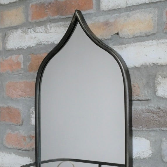 Close up of an arched wall mirror with a dark metal frame, shown against a rustic brick wall background.