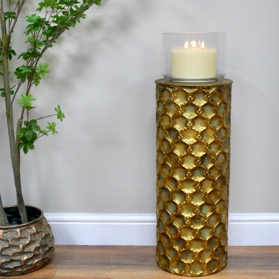 A tall cylindrical gold pedestal with a textured, scale-like pattern holds a lit white pillar candle in a clear glass holder, set against a neutral grey wall with a wooden floor and a potted plant on the left.