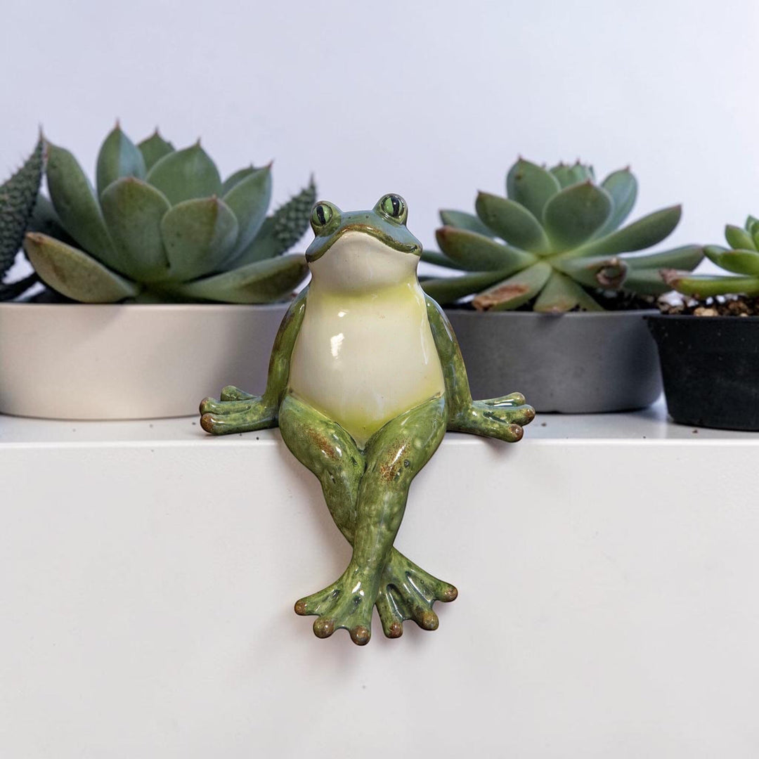 Green polyresin frog shelf sitter with crossed legs sitting on a white ledge with succulent plants in the background