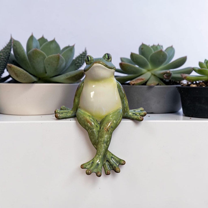Green polyresin frog shelf sitter with crossed legs sitting on a white ledge with succulent plants in the background