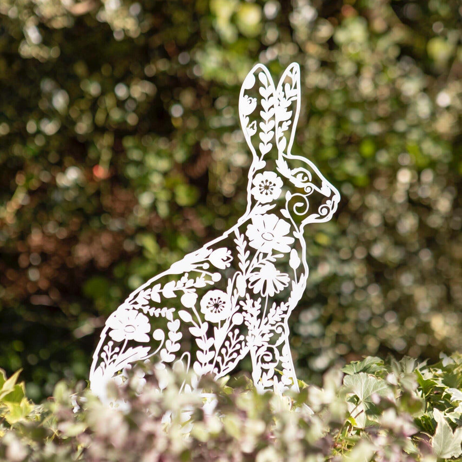 white metal rabbit with cut out flower pattern sitting in a green garden
