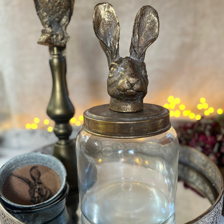 A clear glass jar with a metal lid topped by a sculpted rabbit head, styled on a tray with warm string lights in the background.