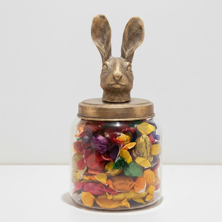 Large clear glass storage jar filled with colorful candies, topped with a distressed golden hare lid.