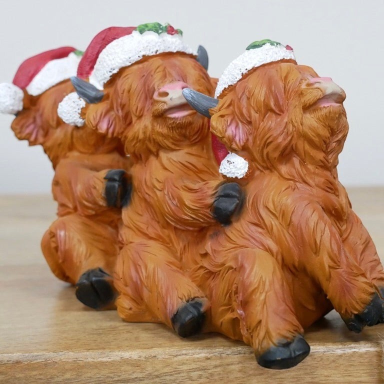 Three small Highland cow figurines wearing Santa hats, arranged in a row on a wooden surface against a plain light background.
