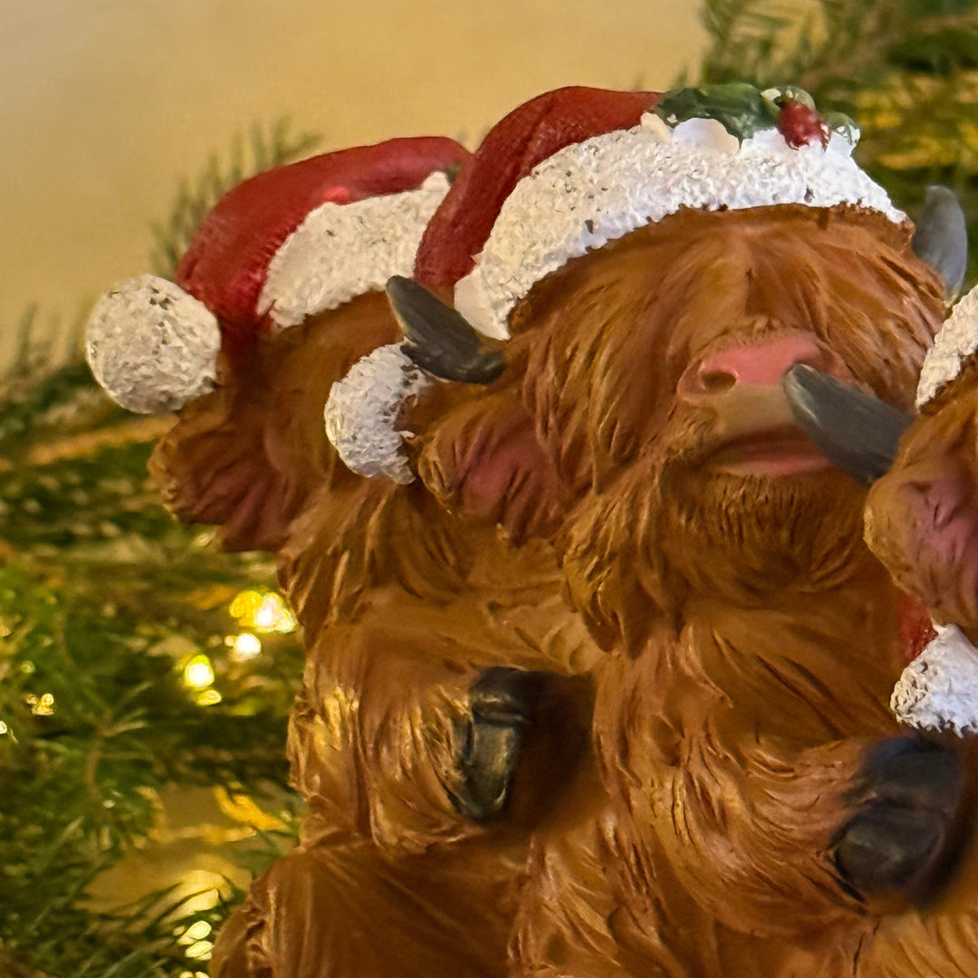 A close-up of a decorative Highland cow figurine wearing a red and white Santa hat, with additional cow figures and green pine branches in the background.