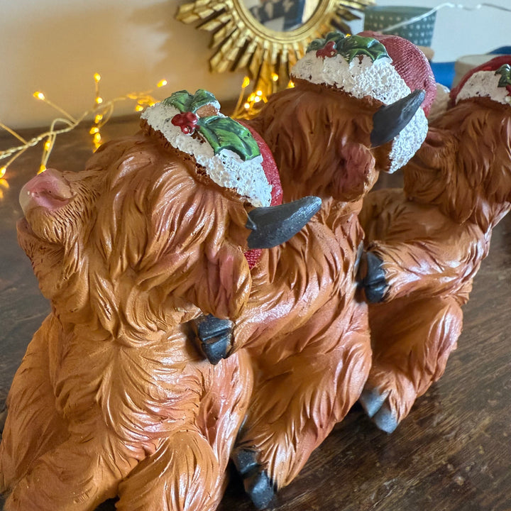 Three small Highland cow figurines with brown fur and red-and-white hats decorated with green leaves and red berries, arranged on a wooden surface.