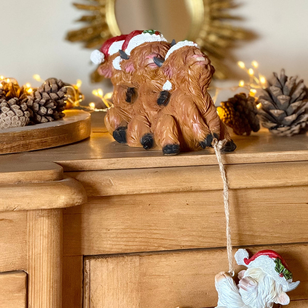 A decorative Christmas ornament shaped like three stacked brown highland cows wearing Santa hats, displayed on a wooden cabinet with pinecones and warm string lights in the background.