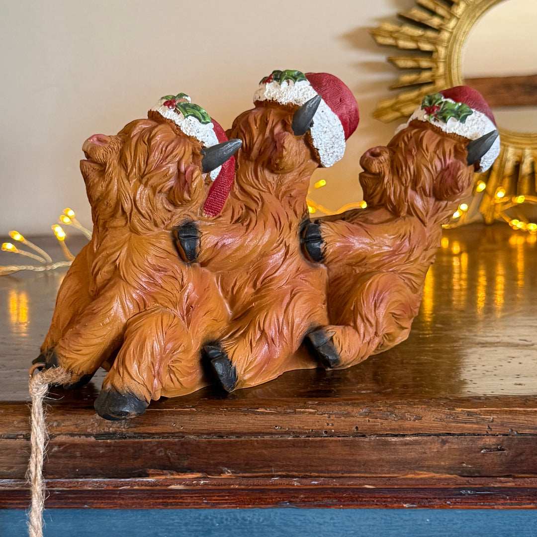 Three small Highland cow figurines posed together on a wooden surface, each wearing a red Santa hat with green and white trim. The scene is lit with warm fairy lights in the background.