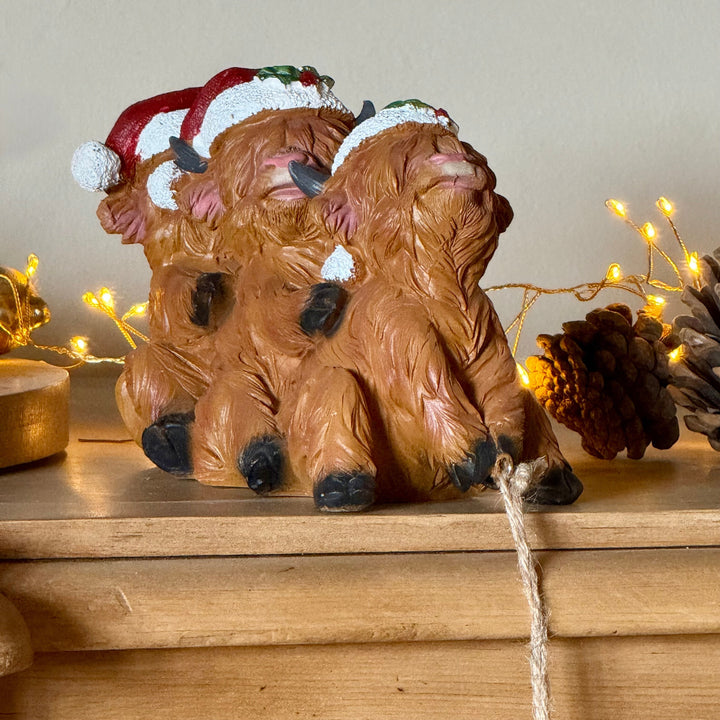 A decorative figurine of three highland cows wearing Santa hats, displayed on a wooden shelf with warm string lights and pinecones in the background.