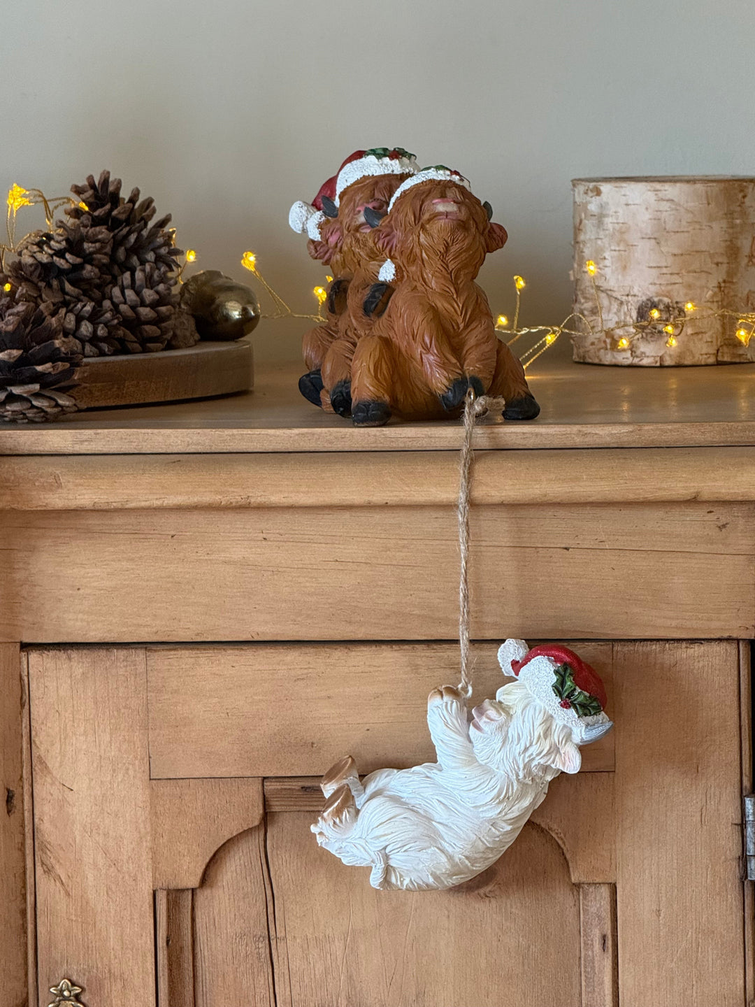 A Christmas ornament with brown highland cows with a white cow hanging from a string, wearing a Santa hat, displayed on a wooden cabinet next to pinecones and warm string lights.