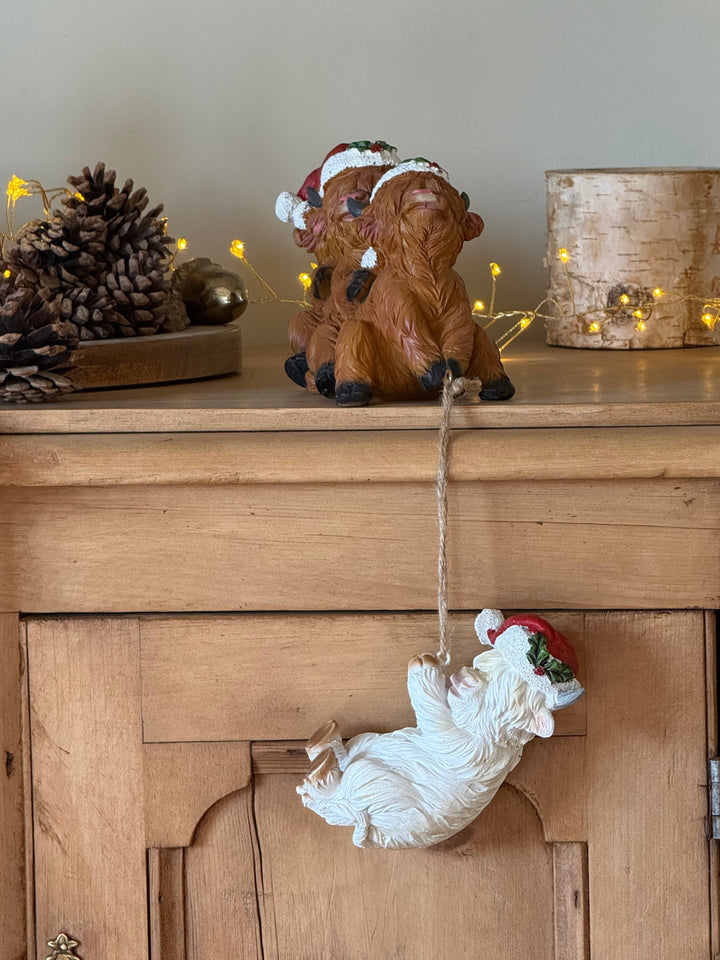 A Christmas ornament with brown highland cows with a white cow hanging from a string, wearing a Santa hat, displayed on a wooden cabinet next to pinecones and warm string lights.