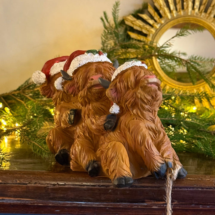 Three small brown Highland cow figurines wearing Santa hats, posed together on a dark wooden surface with holiday greenery and warm lights in the background.