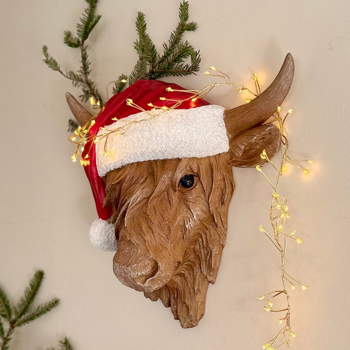 Decorative cow head wearing a Santa hat with string lights on a wall.