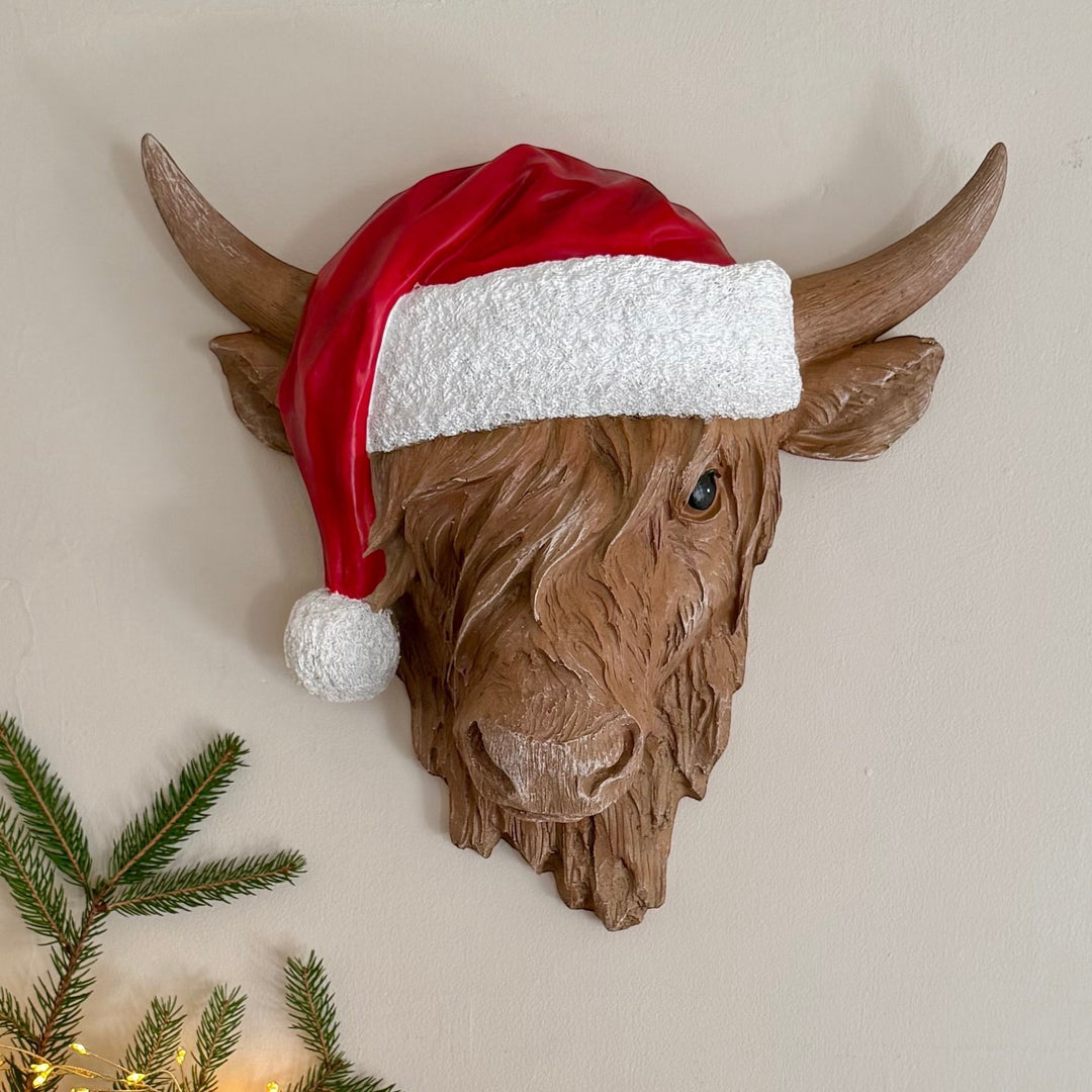 Resin highland cow head wall hanger wearing a red Santa hat, hung on a neutral wall with pine foliage and fairy lights below