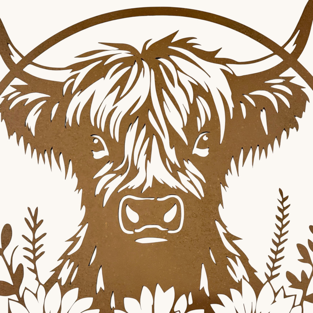 A close-up of a brown laser-cut metal wall art piece featuring a Highland cow head with long horns and decorative foliage, shown against a light off-white background.